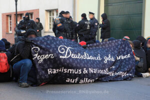 Eine Gruppe von Personen sitzt vor einer Reihe Polizist*innen und hält ein Transparent.