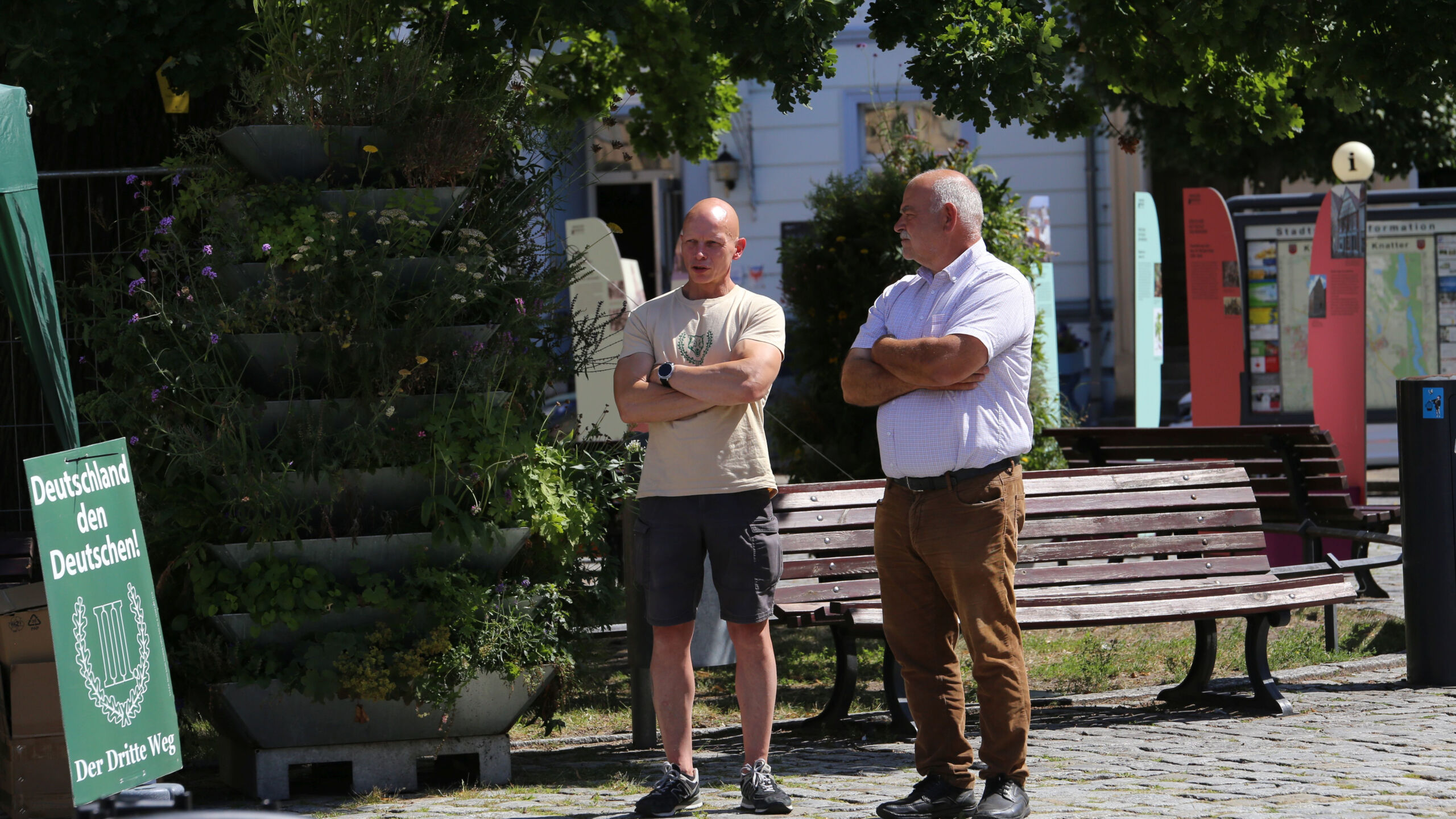 Zwei Personen stehen mit verschränkten Armen vor einer Parkbank